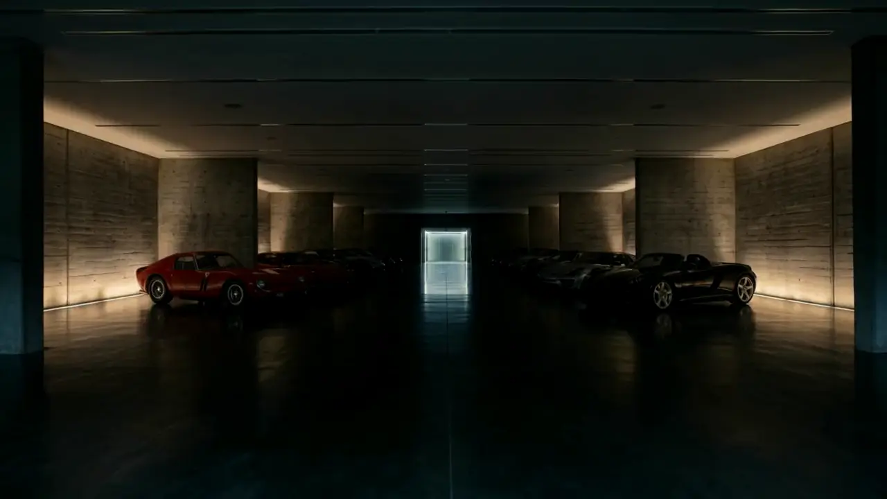 A private underground car gallery below a house, with cars parked in a lit aisle under a concrete ceiling